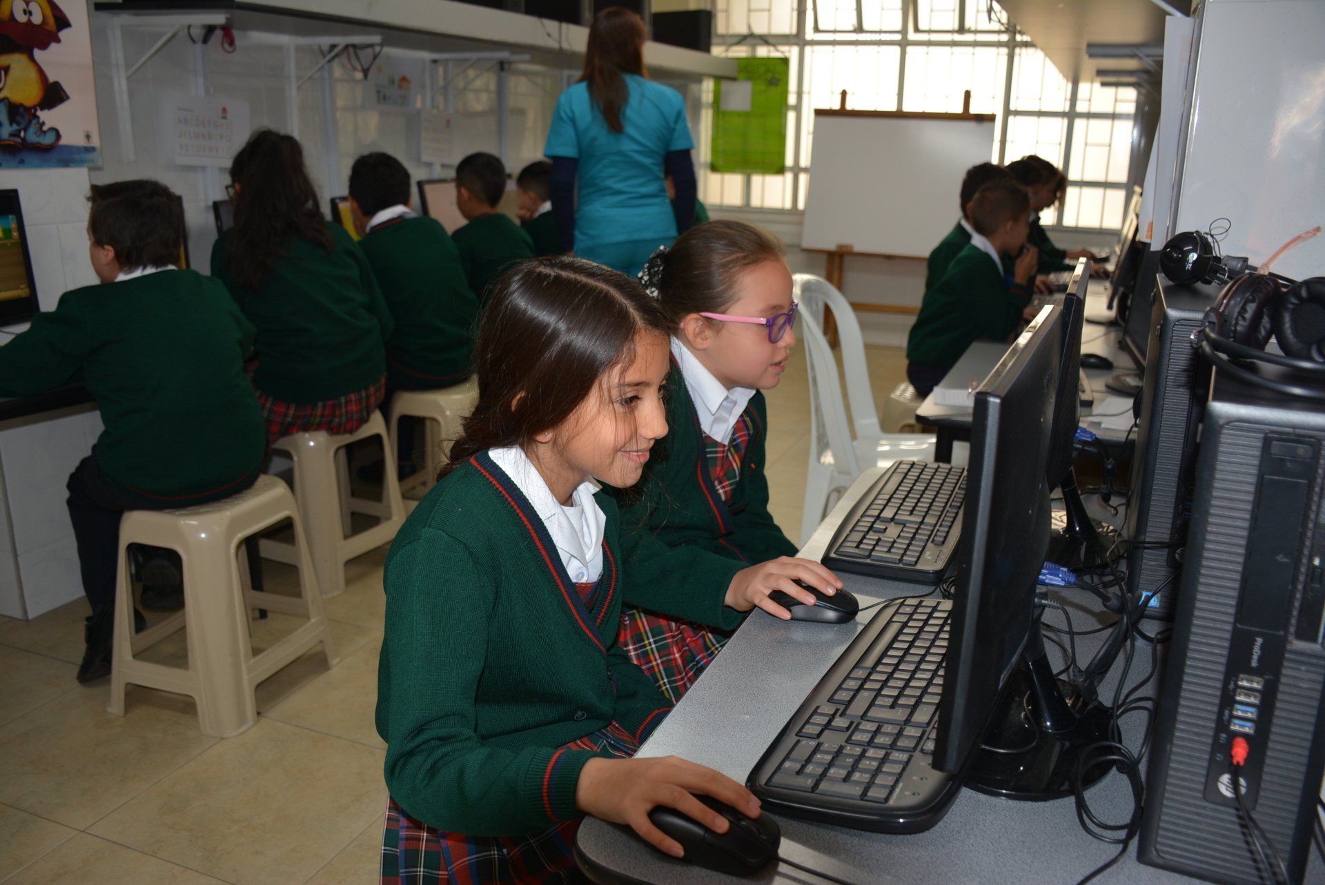 Estudiantes usando computadoras de escritorio en un laboratorio de computación de un aula, con un profesor cerca.