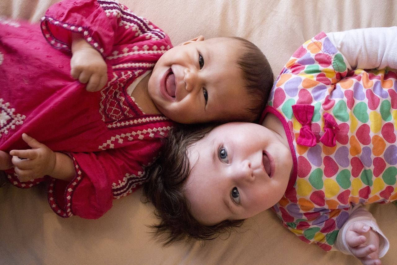 Two babies lie head-to-head on a light surface; one laughs wearing a pink dress, the other in a colorful heart-print top.