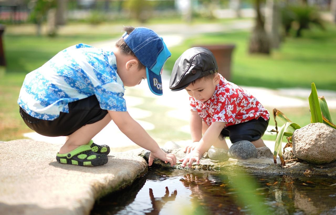 Two children wearing hats and patterned shirts squat beside a pond, reaching their hands into the water in a garden.