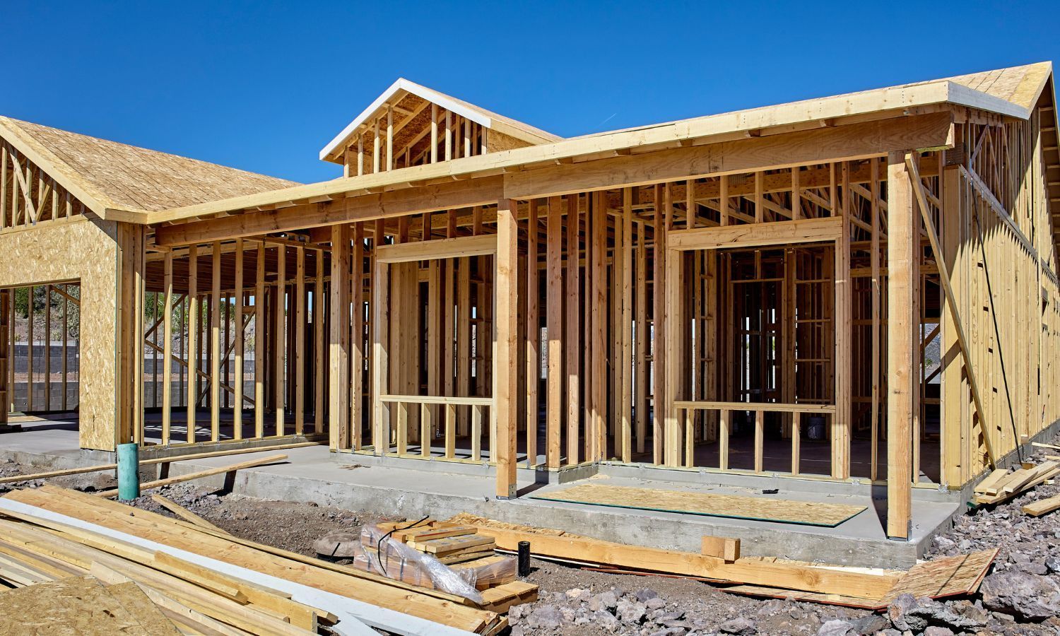 A house frame under construction against a clear blue sky, with wood beams and studs visible.