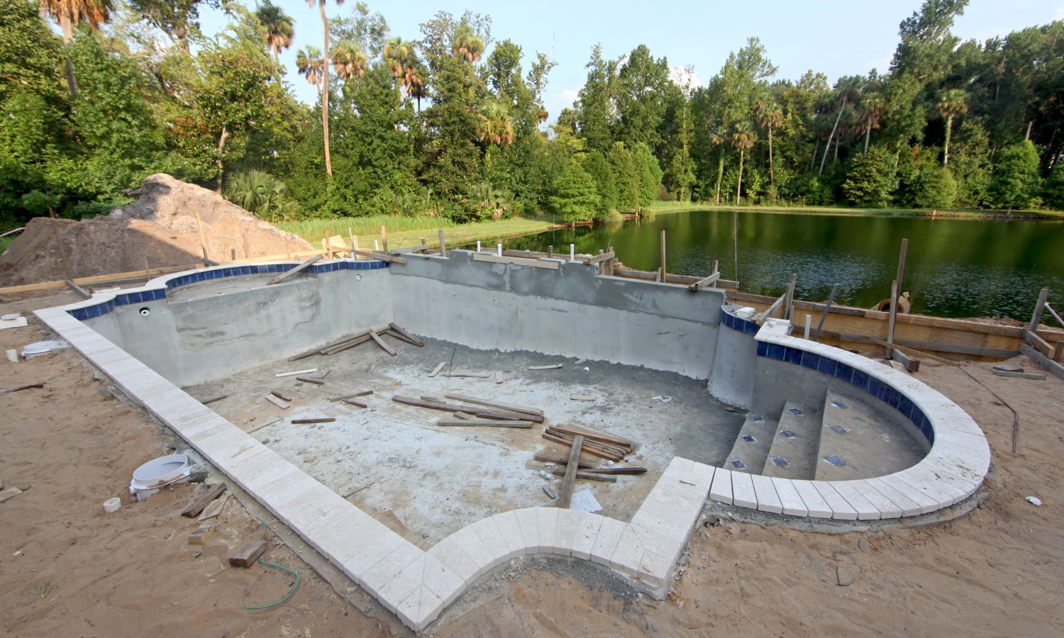 Construction of an empty swimming pool with steps, overlooking a pond, surrounded by trees.