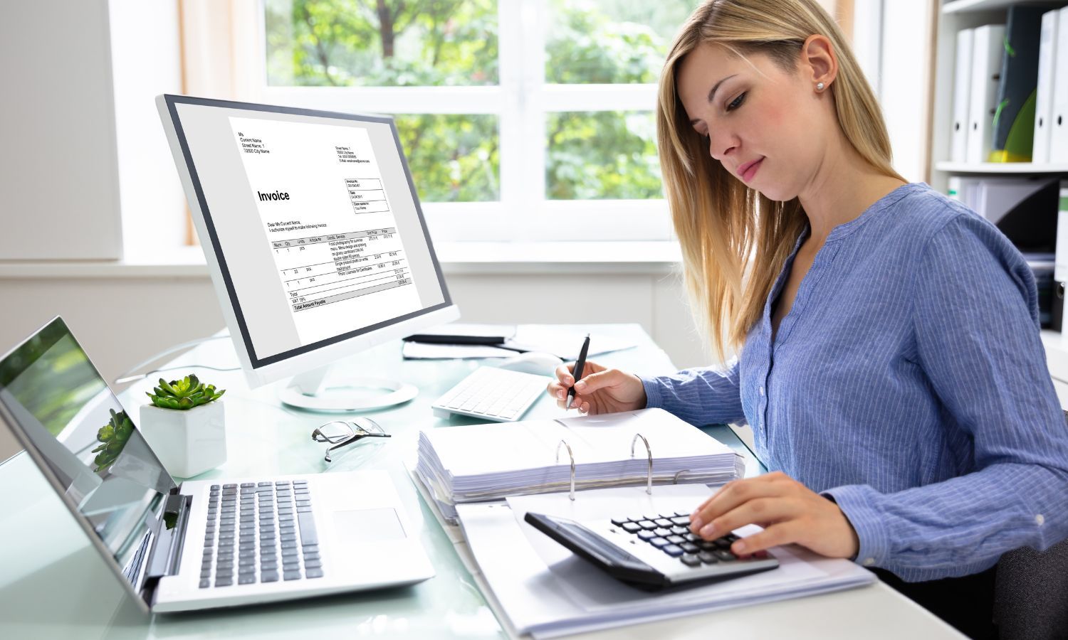 Woman working at a desk with a computer, laptop, calculator, and papers.