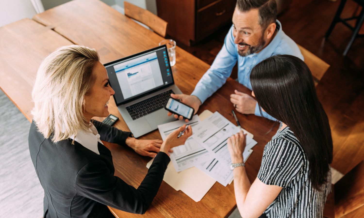 Financial advisor showing paperwork to a couple at a wooden table; laptop and phone present, smiles, office setting.