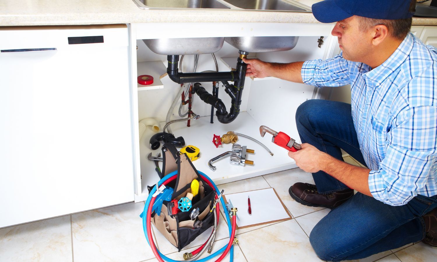 Plumber kneels under a kitchen sink, fixing pipes with a wrench and tools.
