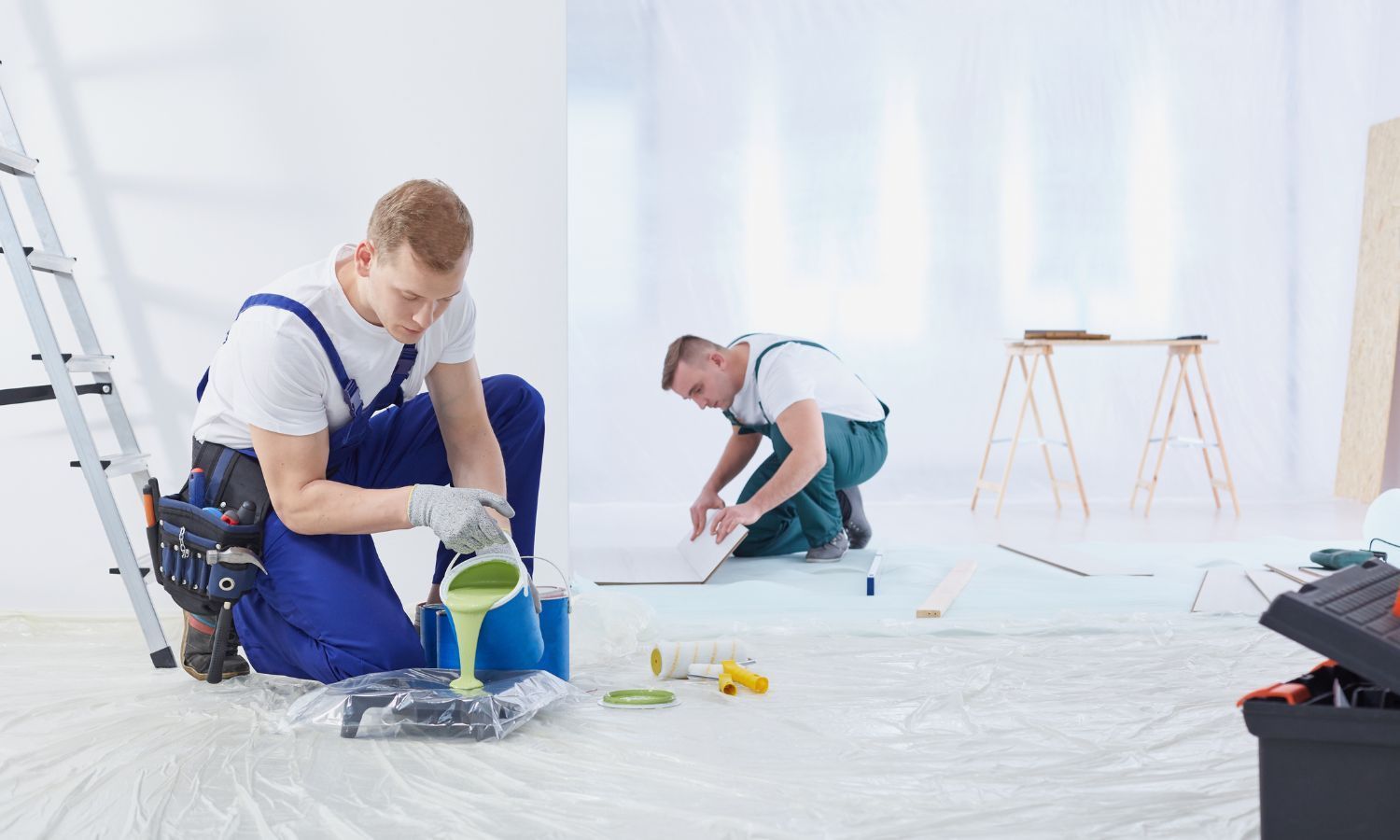 Two painters in a room. One pours green paint into a tray, the other works on the floor.