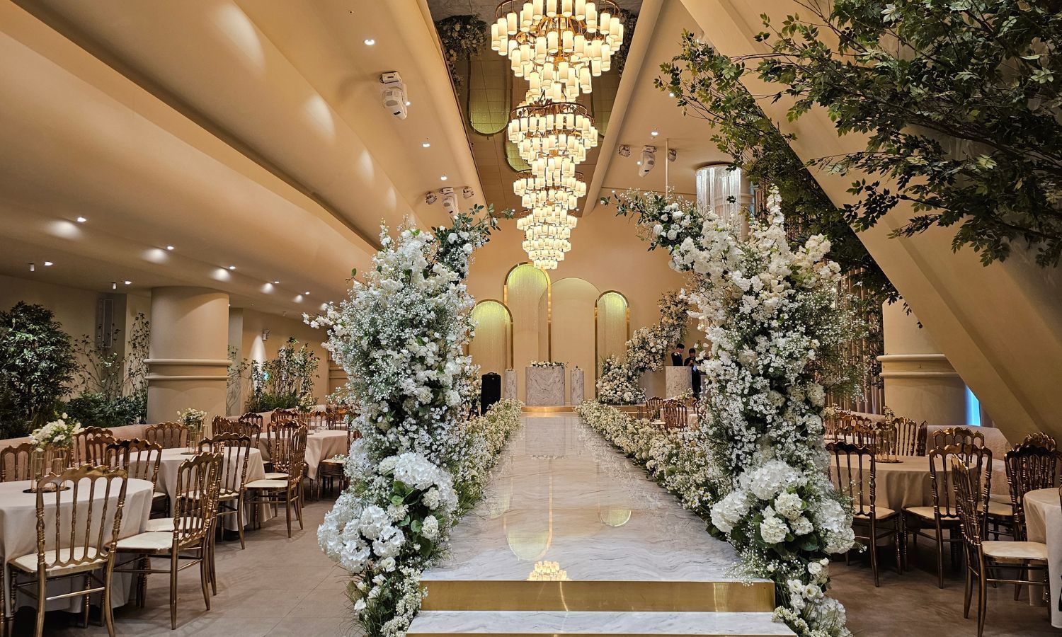 Wedding reception hall decorated with white flowers, gold chairs, and a long aisle.