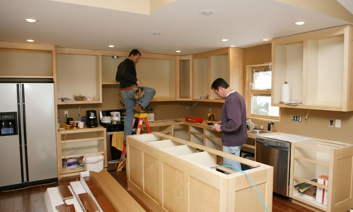 Two men install kitchen cabinets in a beige-walled room. One stands on a ladder.