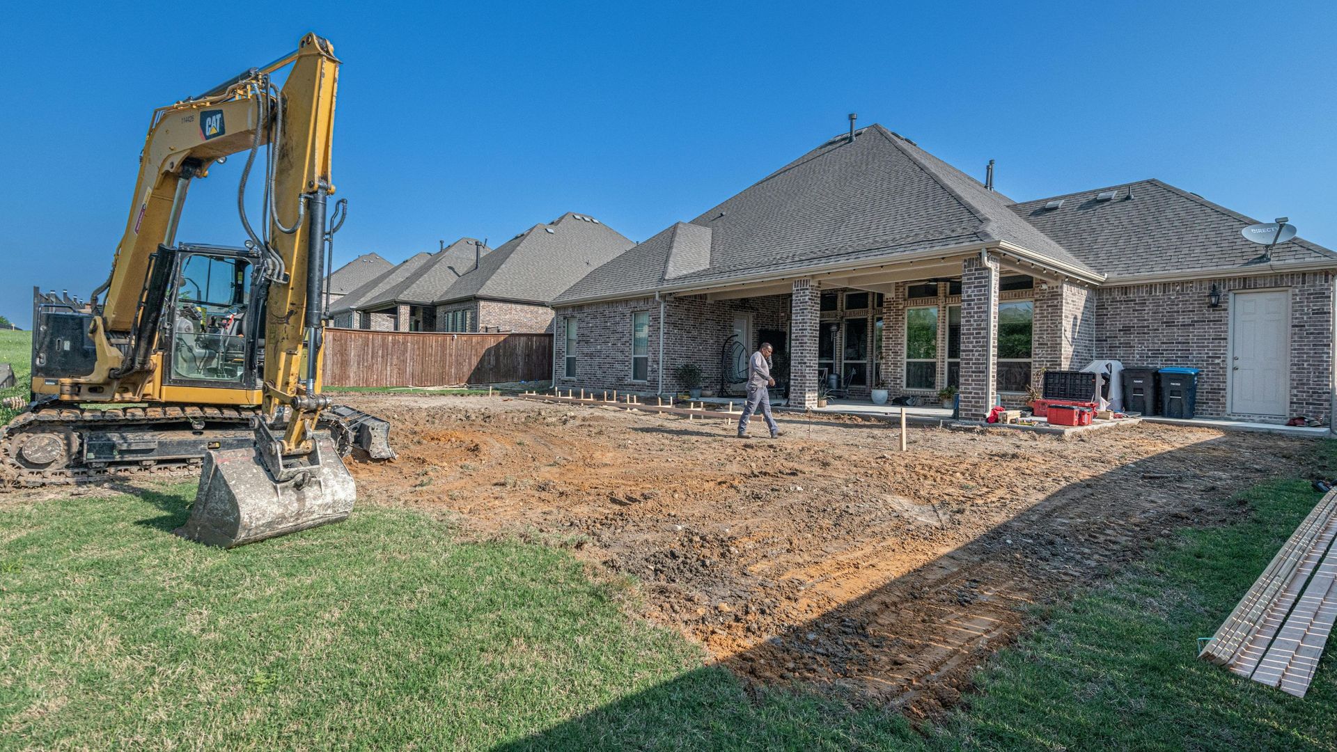 Excavator digging in a backyard, preparing for construction near a brick house on a sunny day.