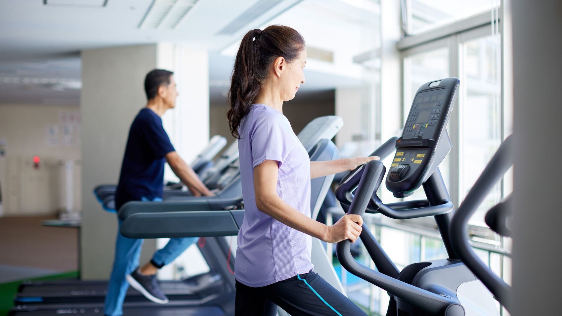 Woman on elliptical machine, man on treadmill at gym near large windows.