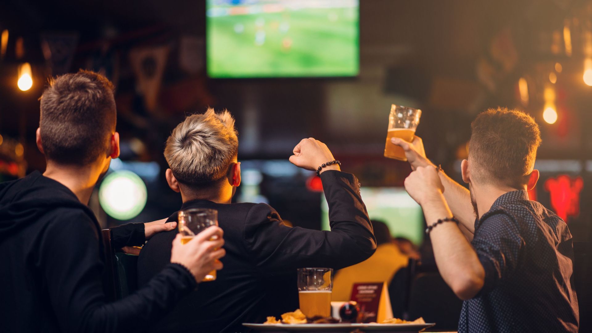 Three men at a bar watch a game on TV, raising beer glasses in celebration. Warm lighting.