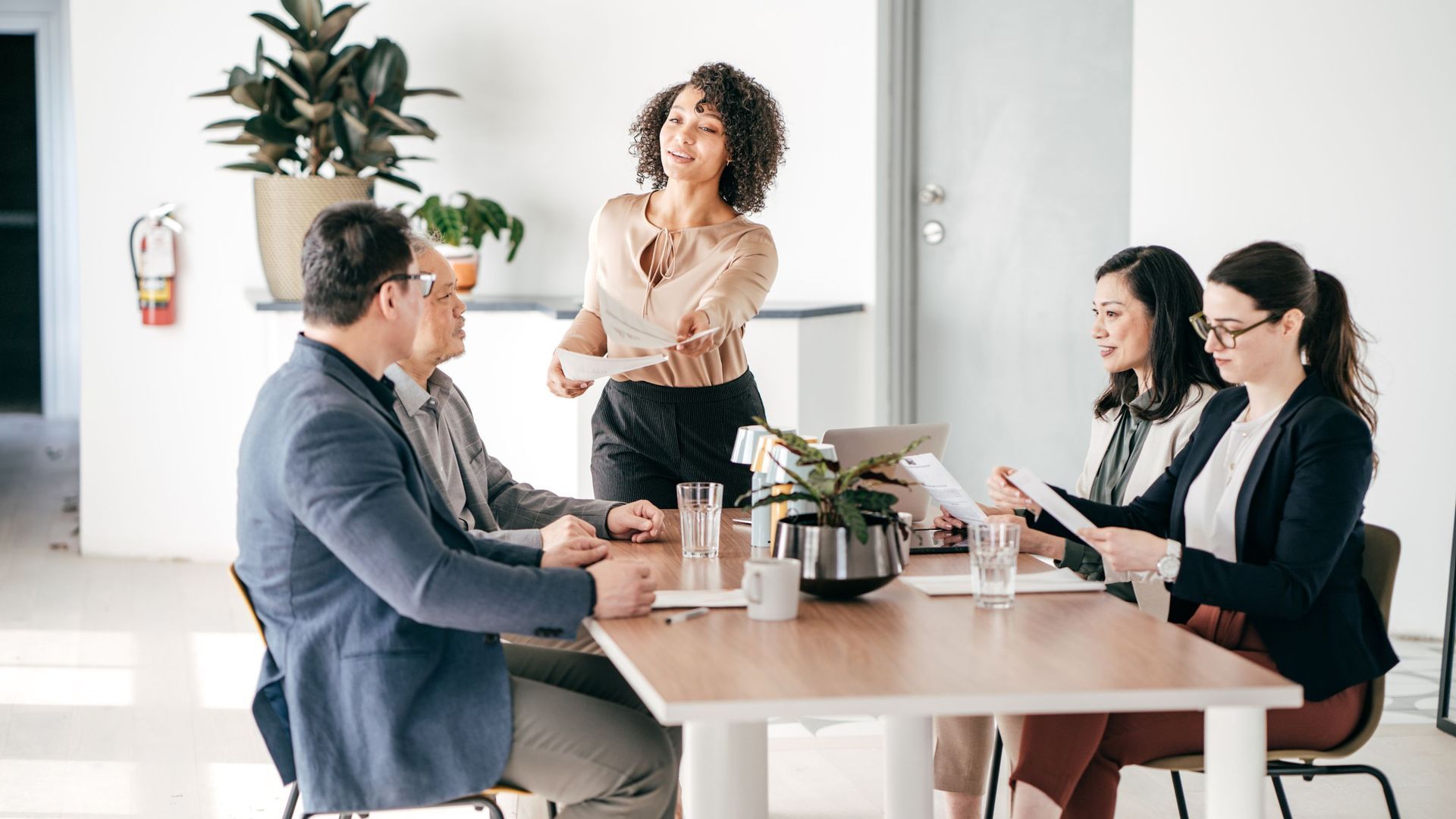Business meeting: Woman hands papers to colleagues around a table. Neutral-toned office with plants.