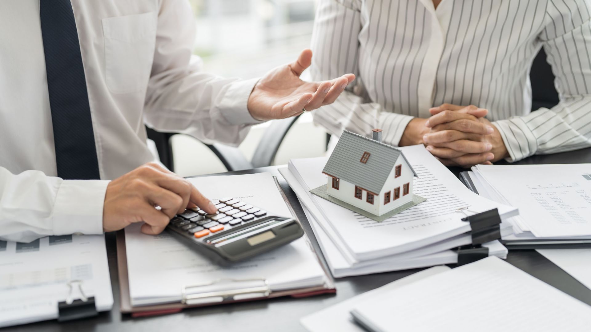 Person using a calculator, pointing with their other hand, while a model house sits on paperwork.
