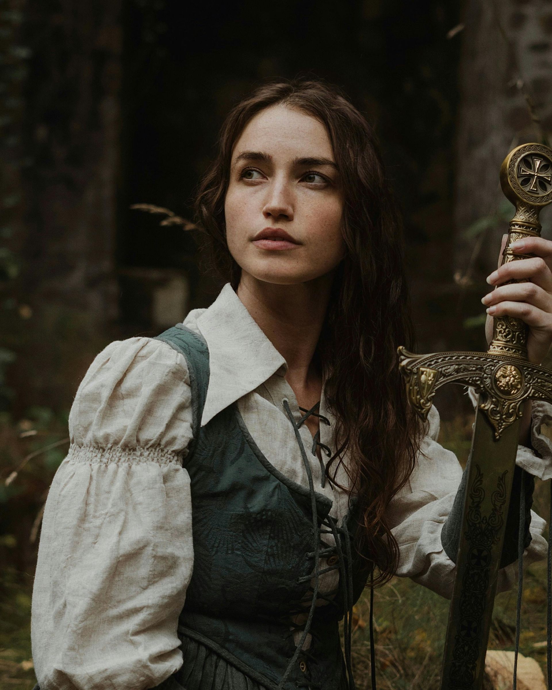 Woman in teal vest and white shirt, holding ornate sword. Forest background.