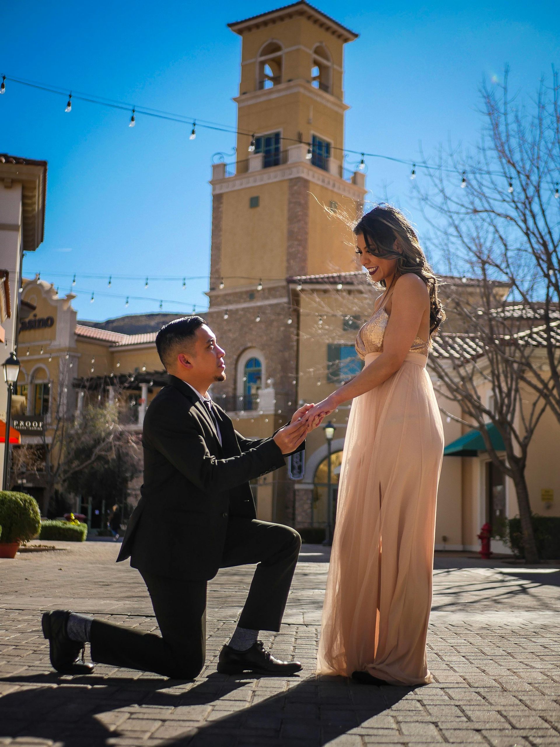 Man kneeling, proposing to a woman. They stand on brick path with buildings and tower in background.
