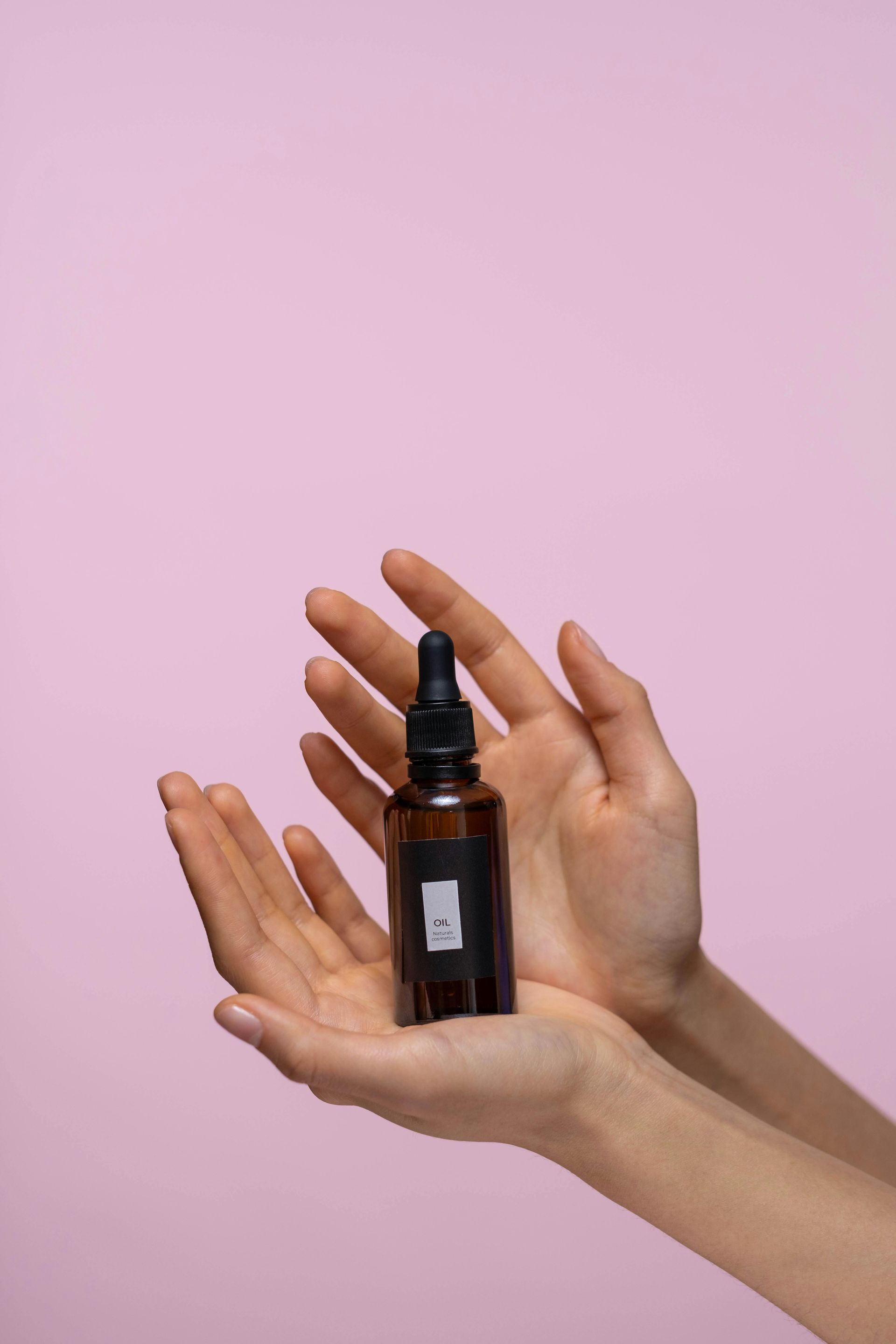 Hands holding a brown dropper bottle on a pink background.