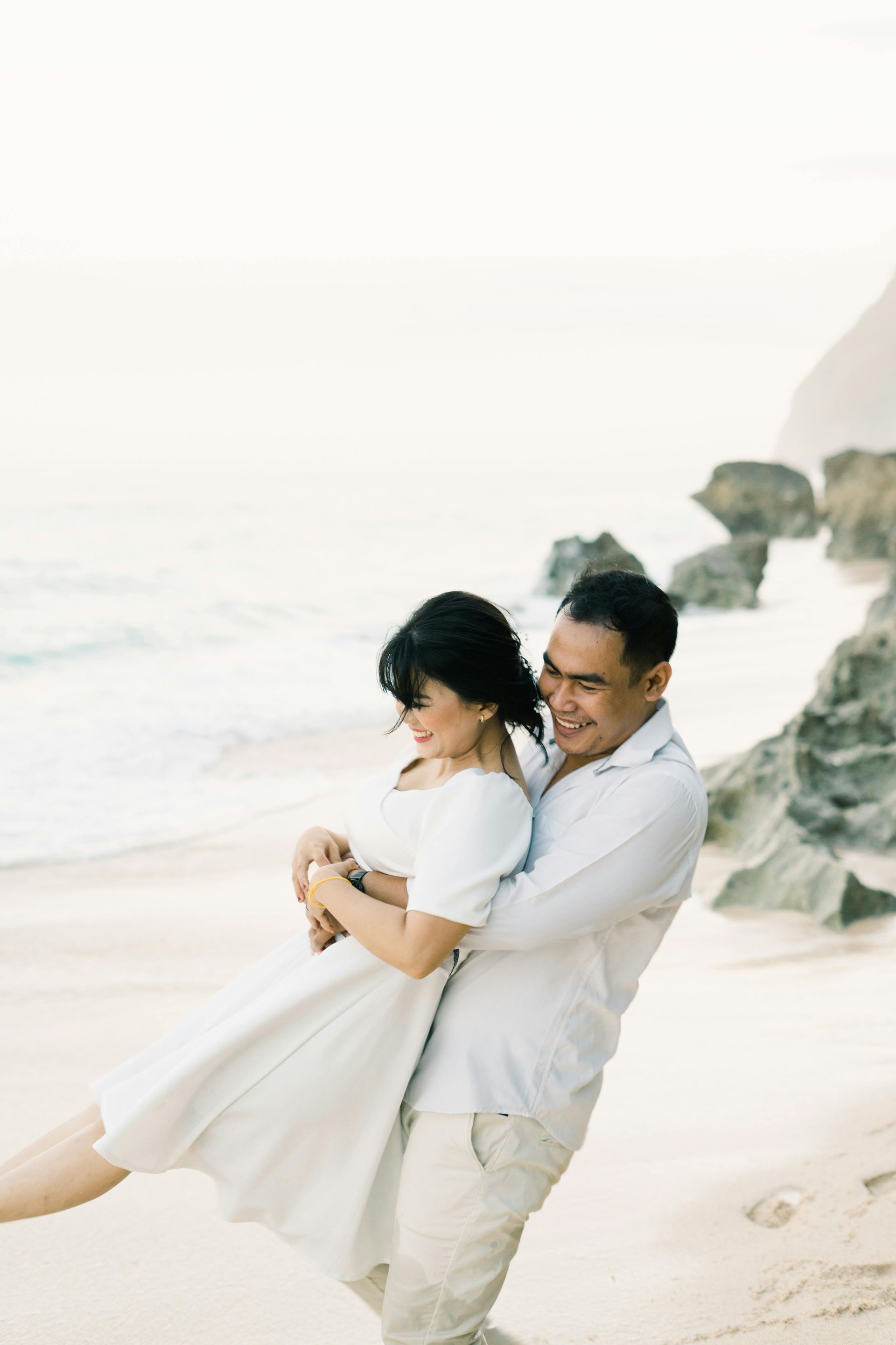 Couple embraces on beach, man spins woman. White clothes, ocean and rocks in background.