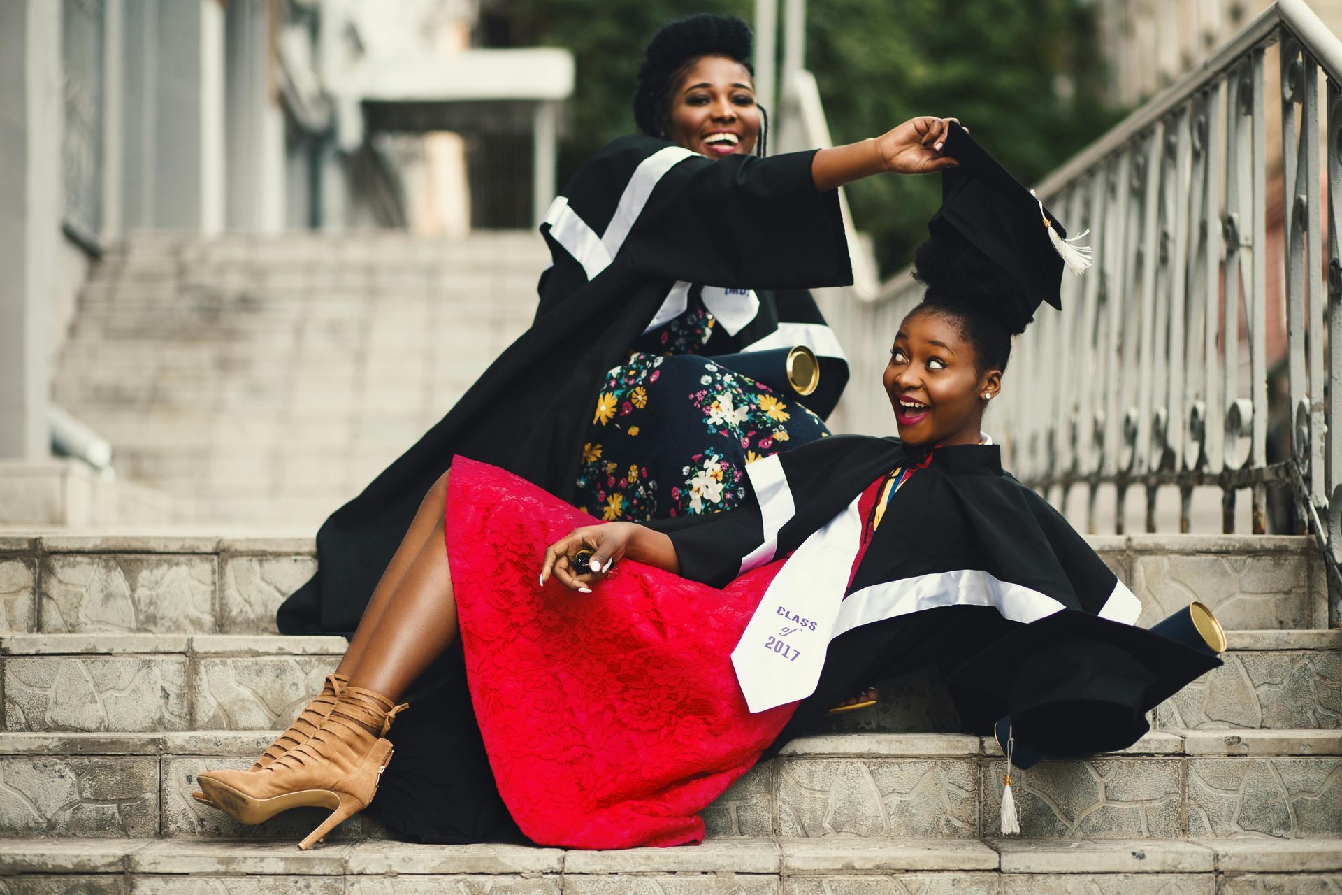 Two graduates in cap and gown, laughing on outdoor steps. One holds the other's mortarboard.