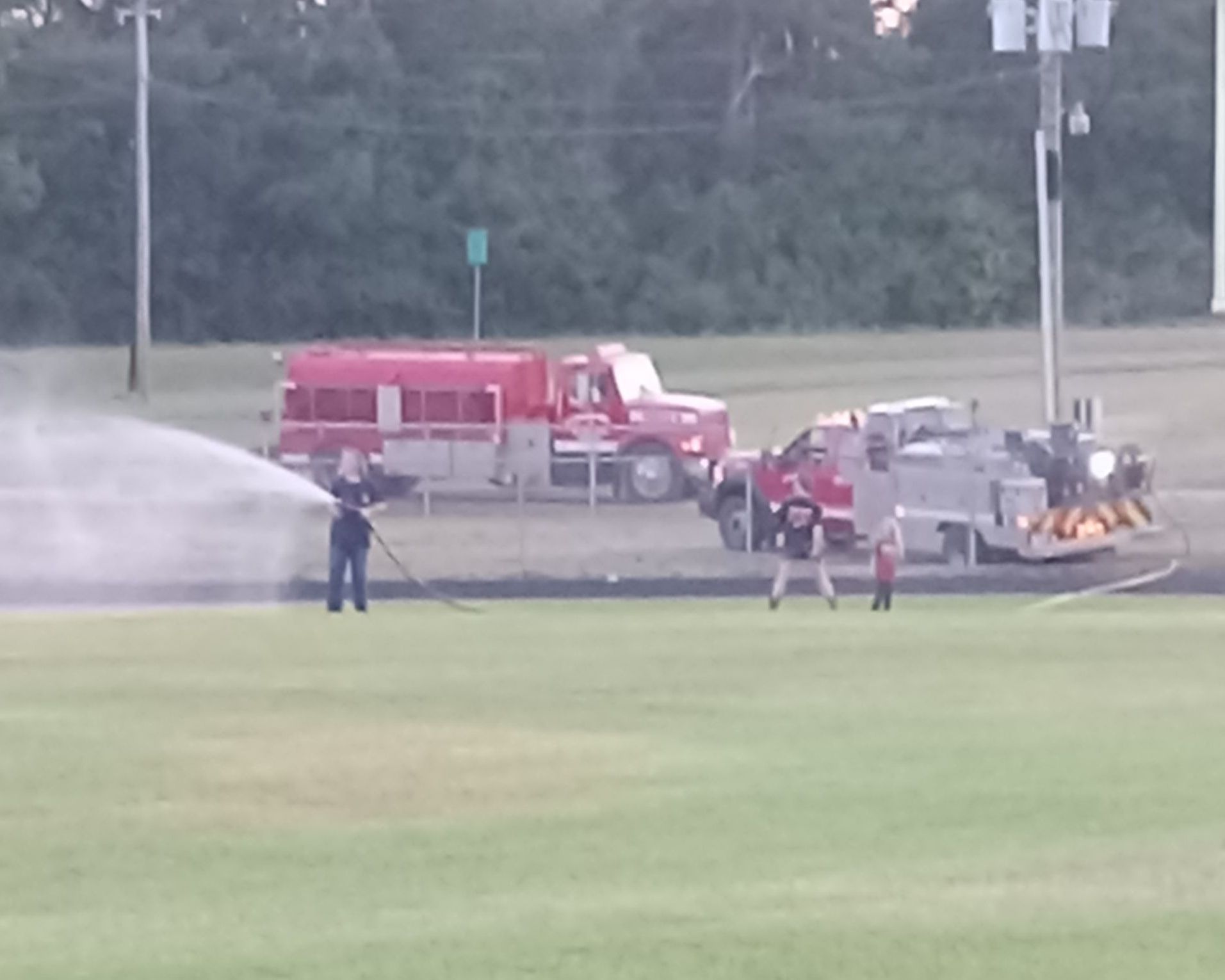 Firefighters spraying water on the ground with two fire trucks nearby. A grassy field is in the foreground.