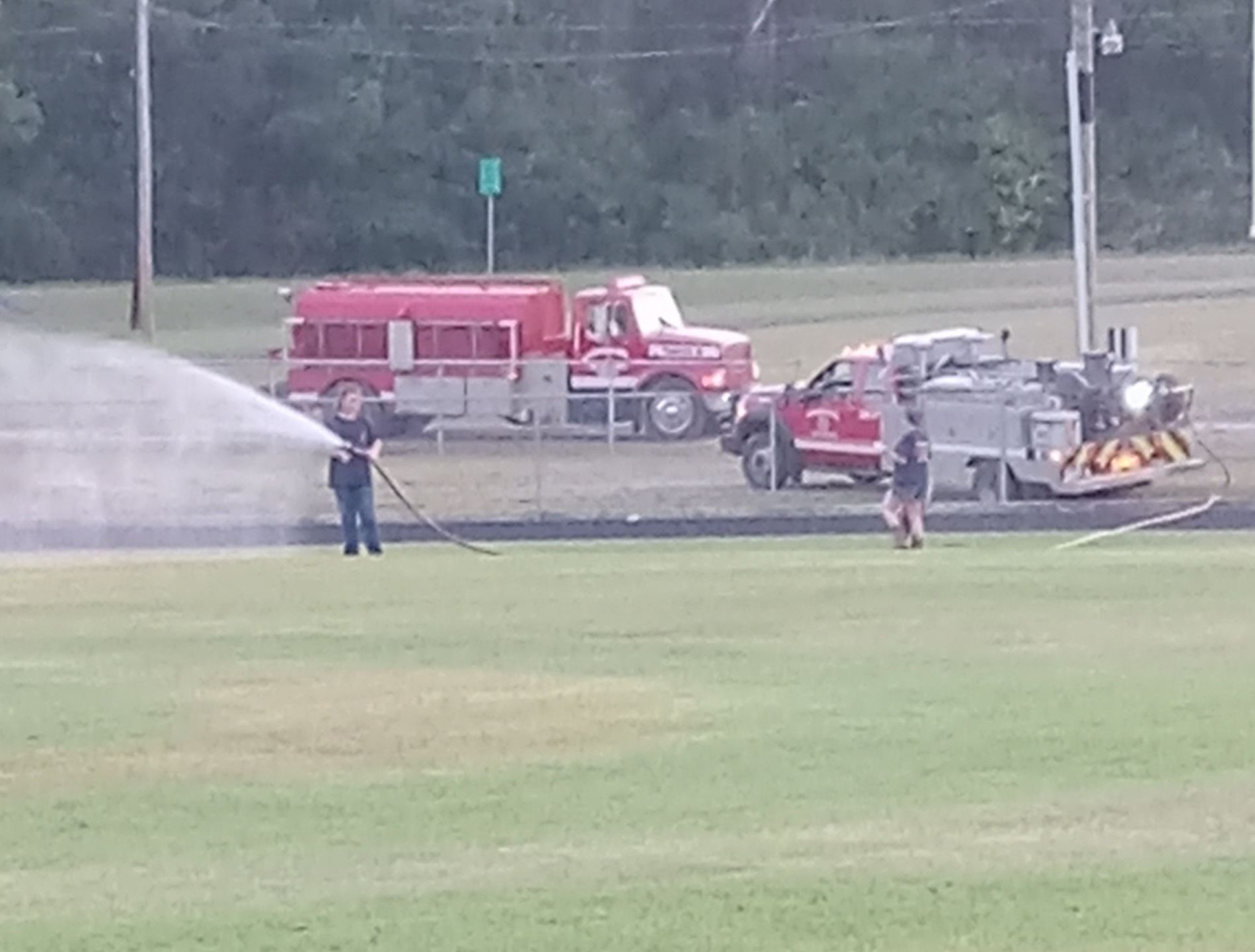 Firefighters spraying water on a grassy field, near two firetrucks and a black track.