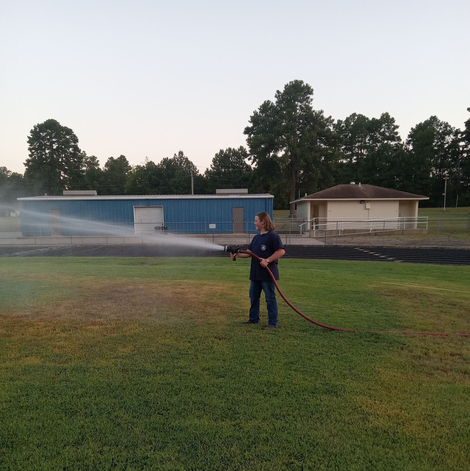 Firefighter spraying water on grass near a blue building at dusk.