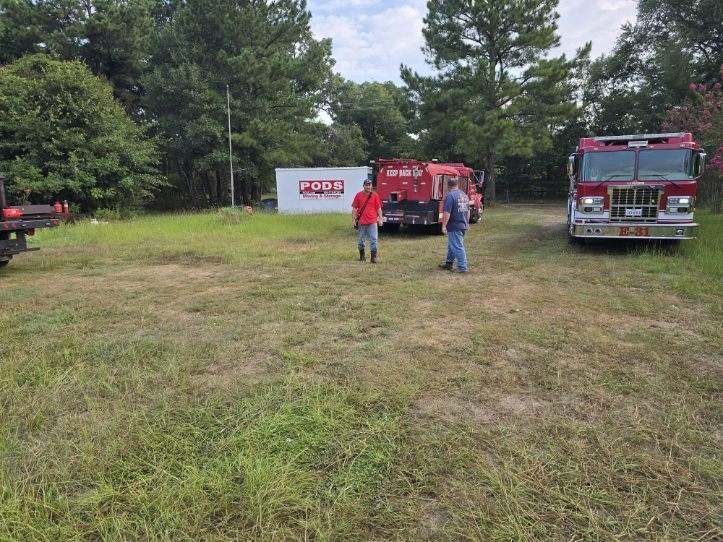 Two men standing near fire trucks in a grassy area, a white building in the background.