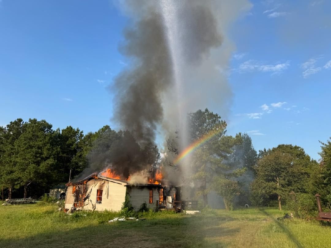 House engulfed in flames, firefighters battling fire with water, with a rainbow appearing in the water stream.