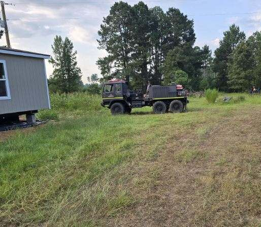 Dark truck parked in grassy field near a building with tall trees in the background.