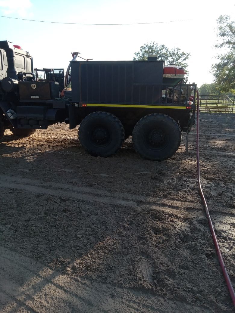 Dark truck with large tires and a water tank, pulling a hose on a dirt field.