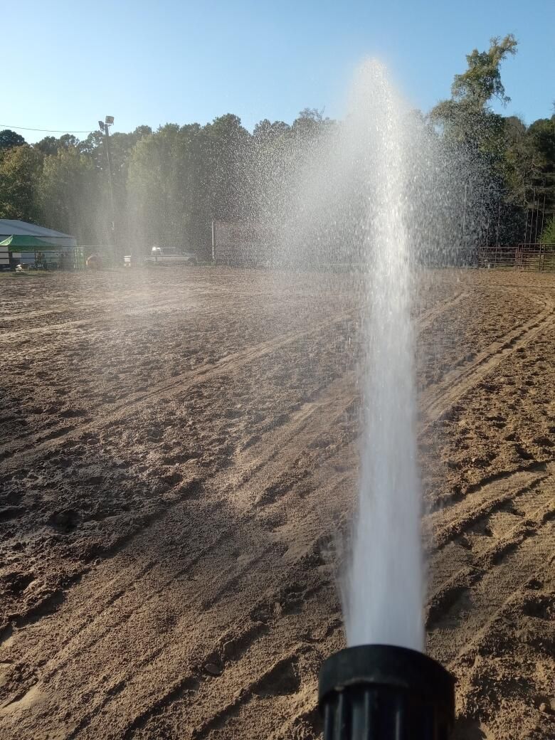 Water spraying from a nozzle irrigating a dry field under a blue sky, with trees in the background.