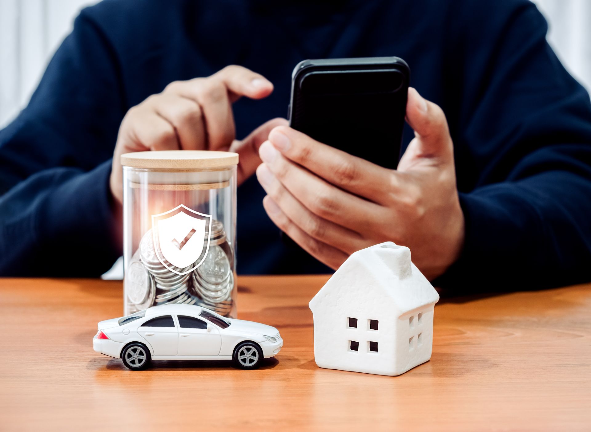 A person is holding a cell phone next to a jar of coins , a car , and a house.