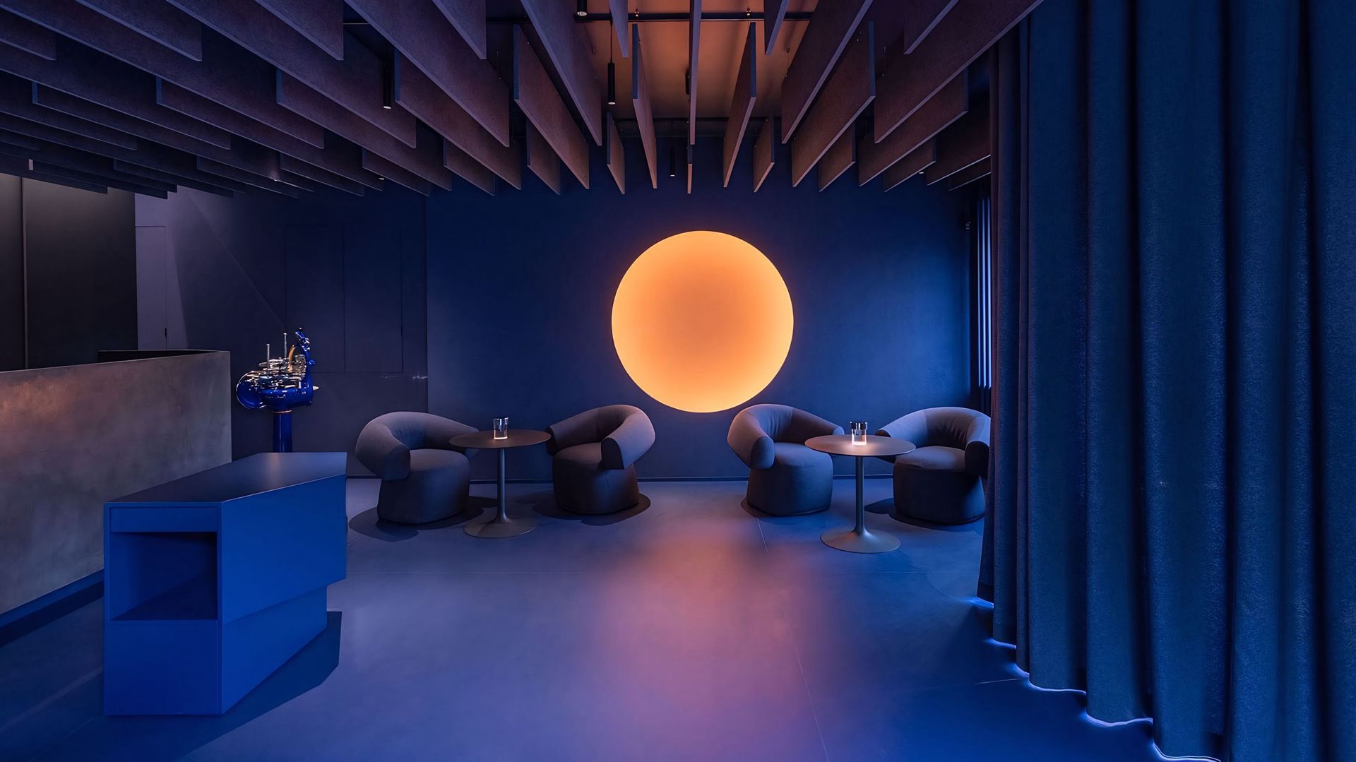Een kamer met blauwe gordijnen, stoelen, tafels en een grote oranje cirkel op de muur.