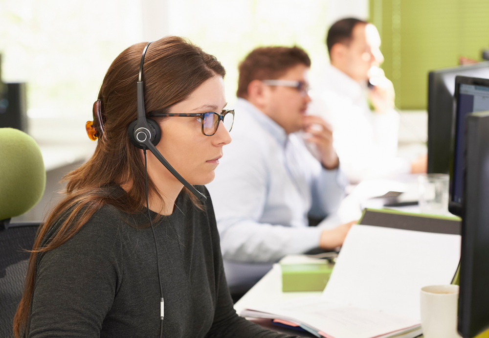A woman wearing a headset is sitting at a desk in front of a computer. A woman wearing a headset is sitting at a desk in front of a computer.