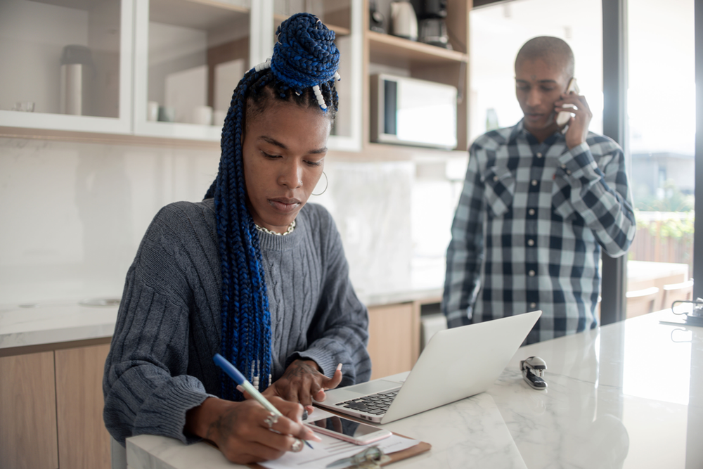 A woman is sitting at a kitchen counter using a laptop computer while a man talks on a cell phone. A woman is sitting at a kitchen counter using a laptop computer while a man talks on a cell phone.