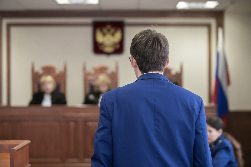 A man in a blue suit is standing in front of a judge in a courtroom.