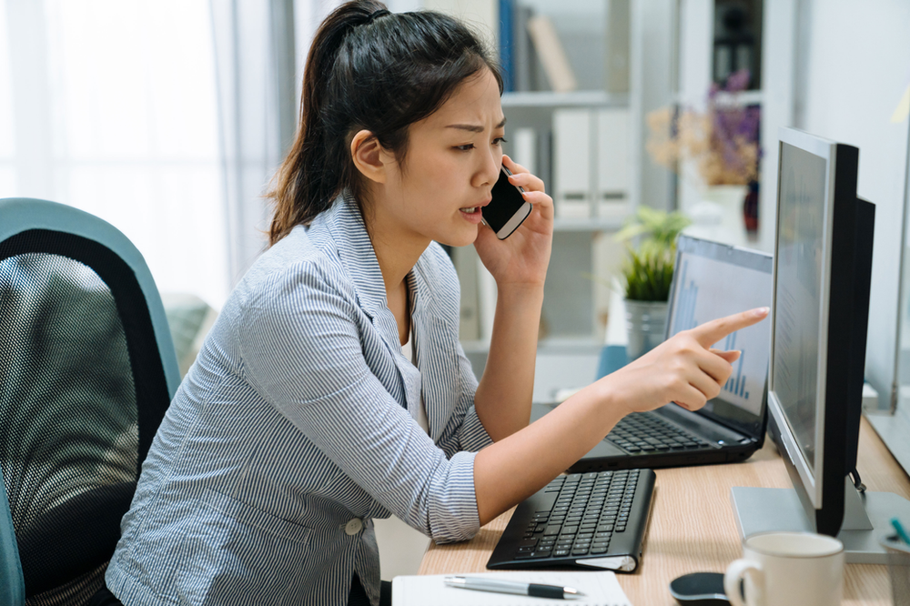 A woman is sitting at a desk talking on a cell phone while pointing at a computer screen. A woman is sitting at a desk talking on a cell phone while pointing at a computer screen.