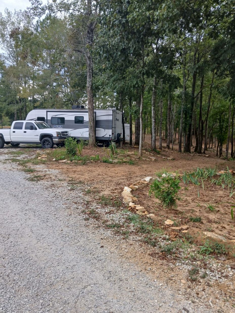 A white pickup truck parked next to a travel trailer on a gravel lot bordered by trees.