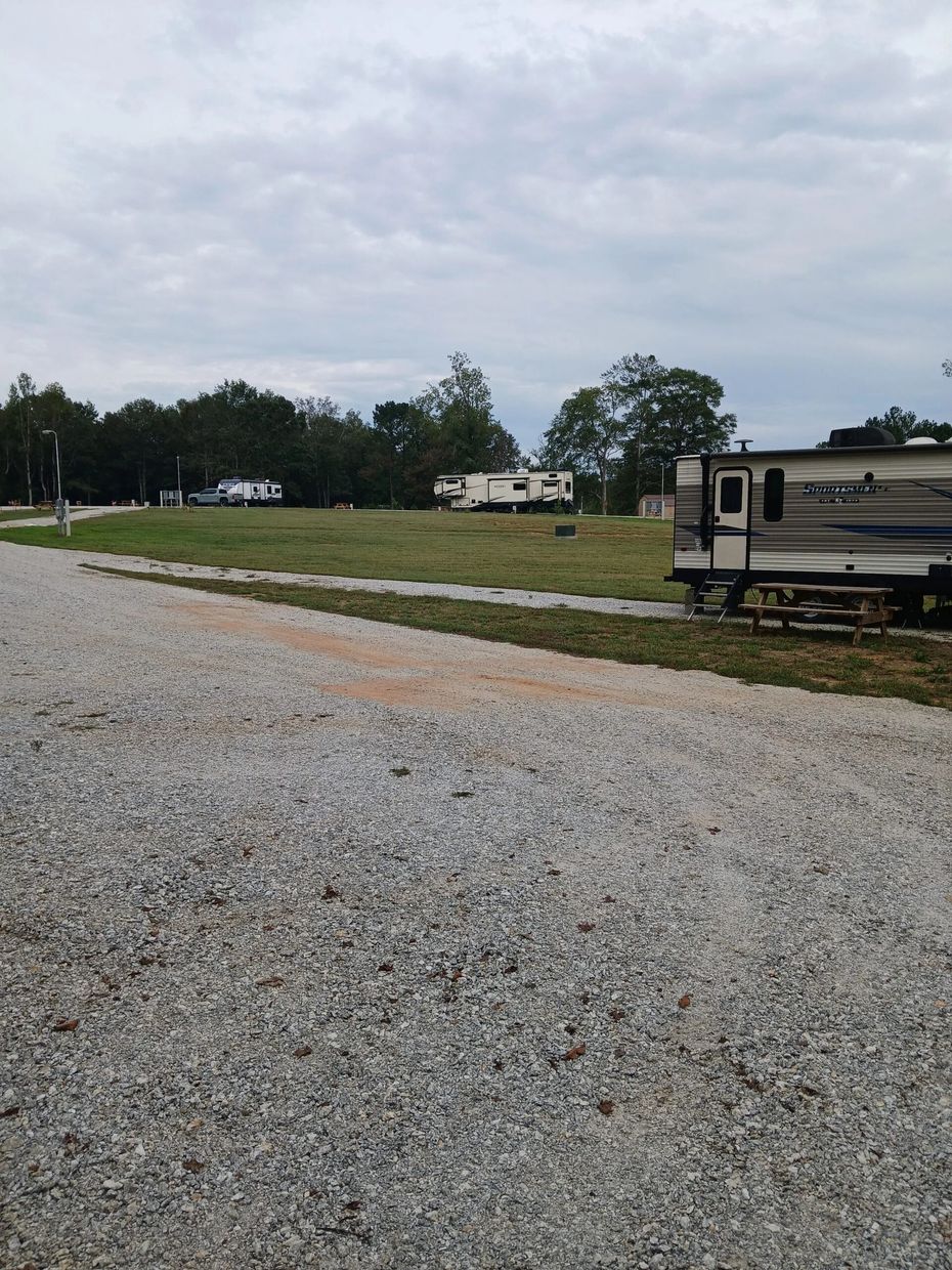 A gravel road leads into a grassy RV park with trailers parked under a cloudy sky.