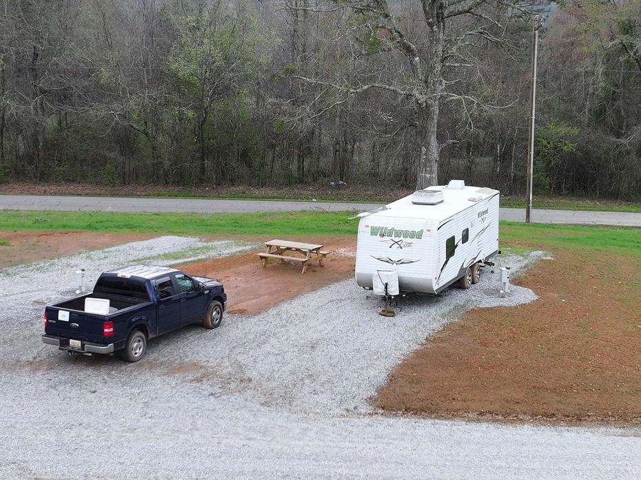 A blue pickup truck parked next to a white Wildwood travel trailer on a gravel campsite near trees and a road.