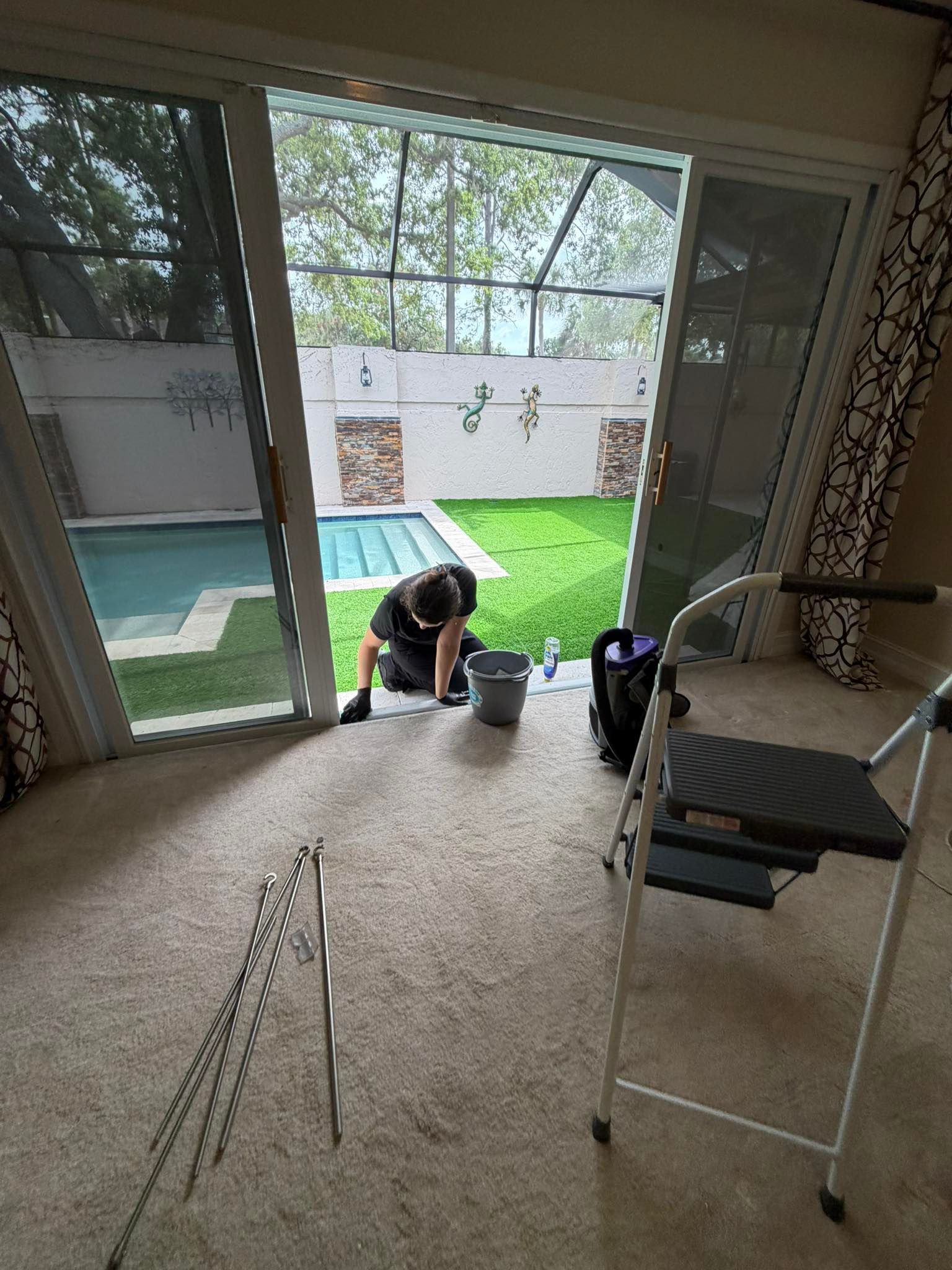 Person kneeling on carpet near sliding doors, working by a patio and pool with tools and a step stool.
