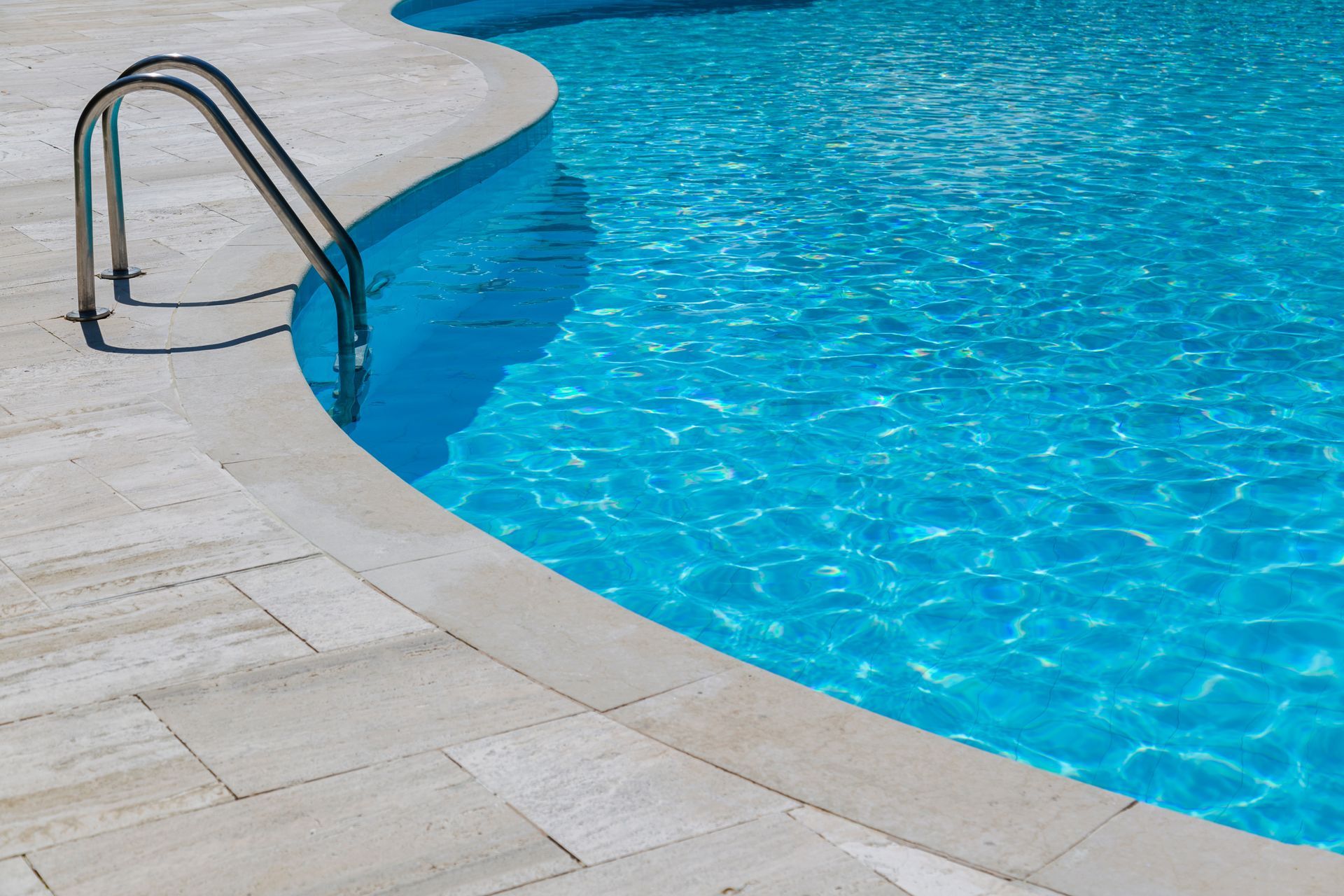 Clear blue swimming pool with a curved edge and metal handrail ladder on a light-colored tiled deck