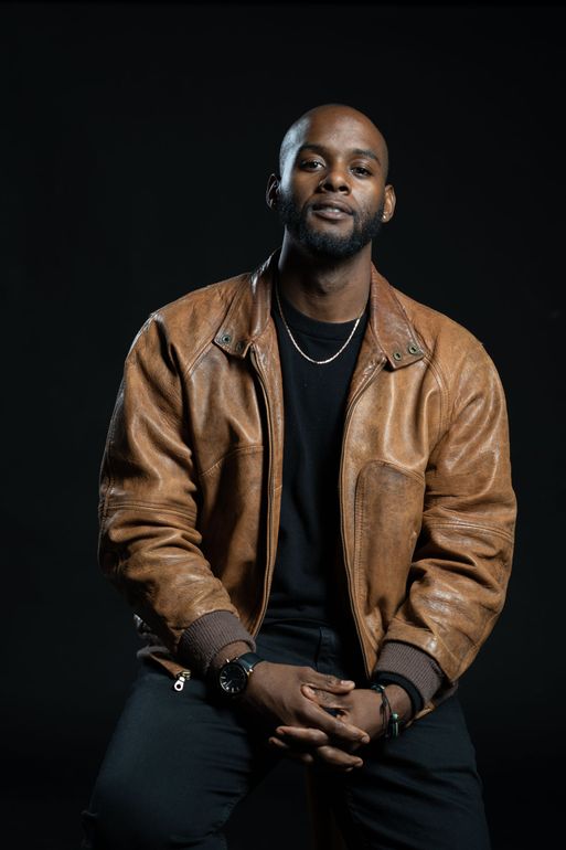 A man with a short beard wearing a brown leather jacket and black shirt, posing against a plain black background.