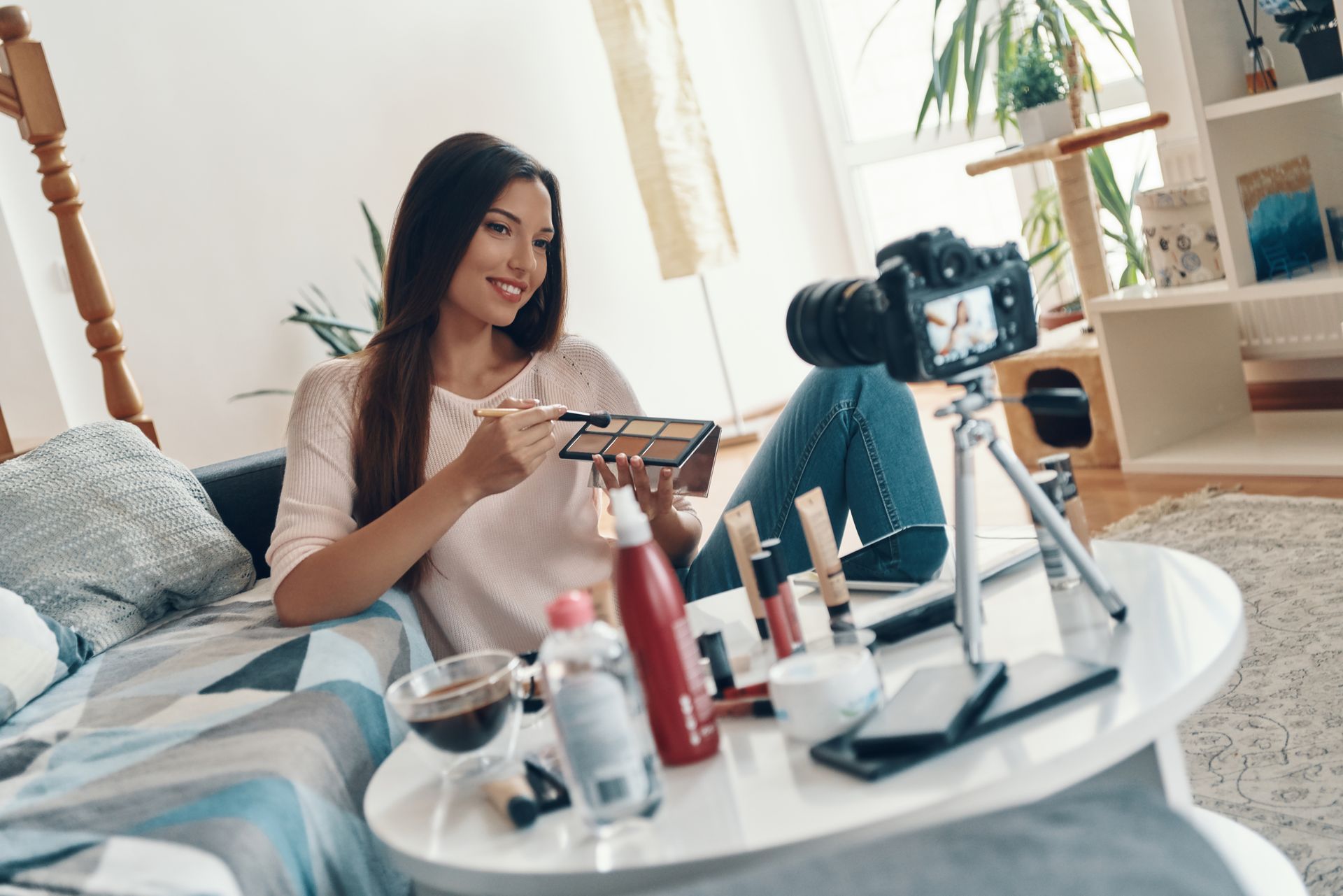 A woman is sitting on a bed applying makeup in front of a camera.