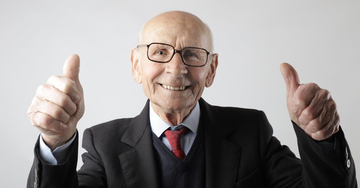 An elderly man in a suit and tie is giving two thumbs up.