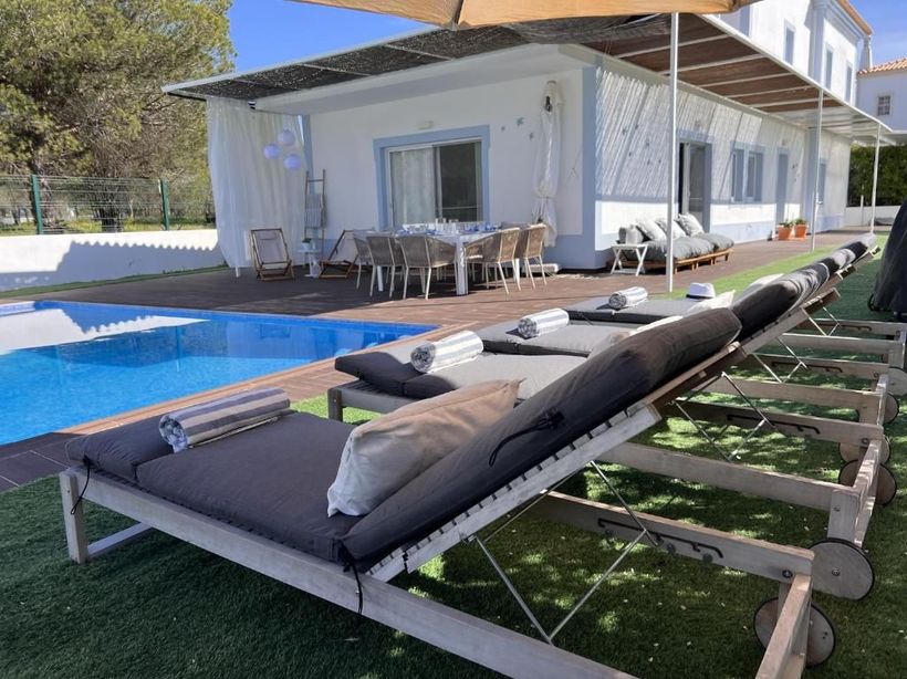 A row of lounge chairs is next to a pool in front of a house.