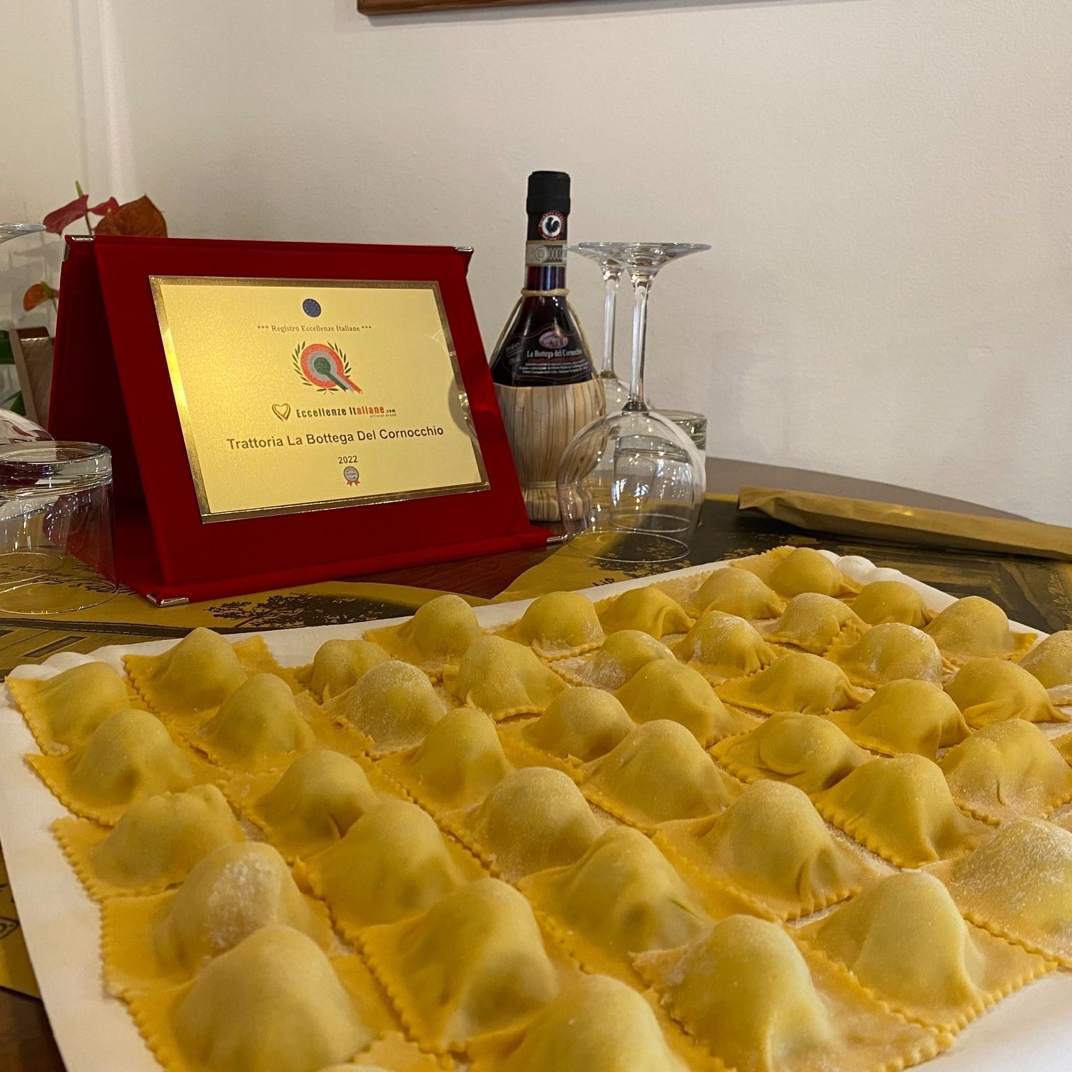 Ravioli freschi su un vassoio, certificato incorniciato, bottiglia di vino e bicchieri su un tavolo.