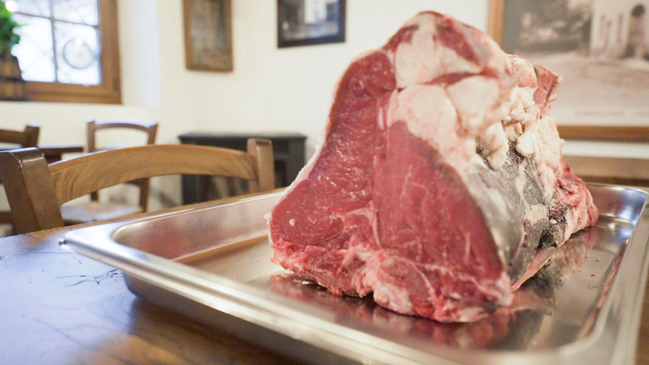 Una grande bistecca di manzo cruda su un vassoio di metallo su un tavolo di legno, in un ambiente da ristorante.