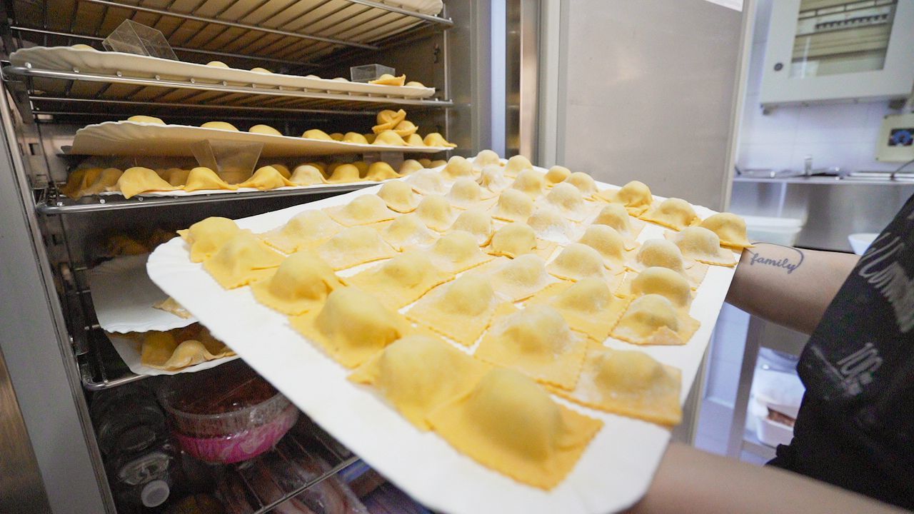 Persona che regge un vassoio di ravioli freschi in una cucina commerciale. Sullo sfondo, scaffali pieni di pasta.