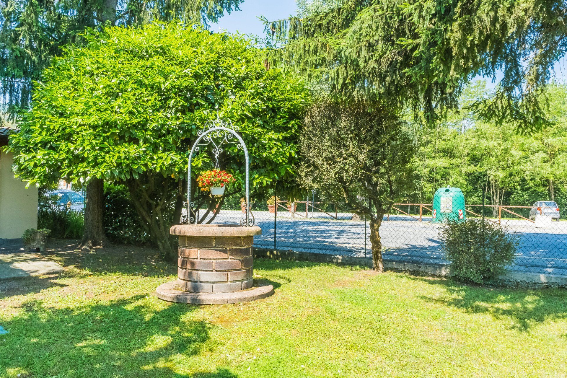 Pozzo in pietra con fiori, in un cortile erboso, circondato da alberi e da un cielo soleggiato.