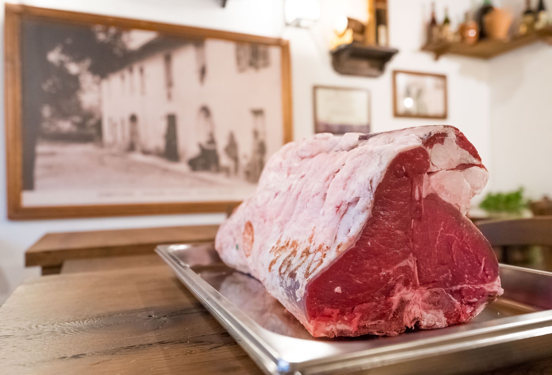 Un grande arrosto di manzo crudo su un vassoio d'argento, posto su un tavolo di legno. Sullo sfondo si vede il dipinto di un edificio in tonalità seppia.
