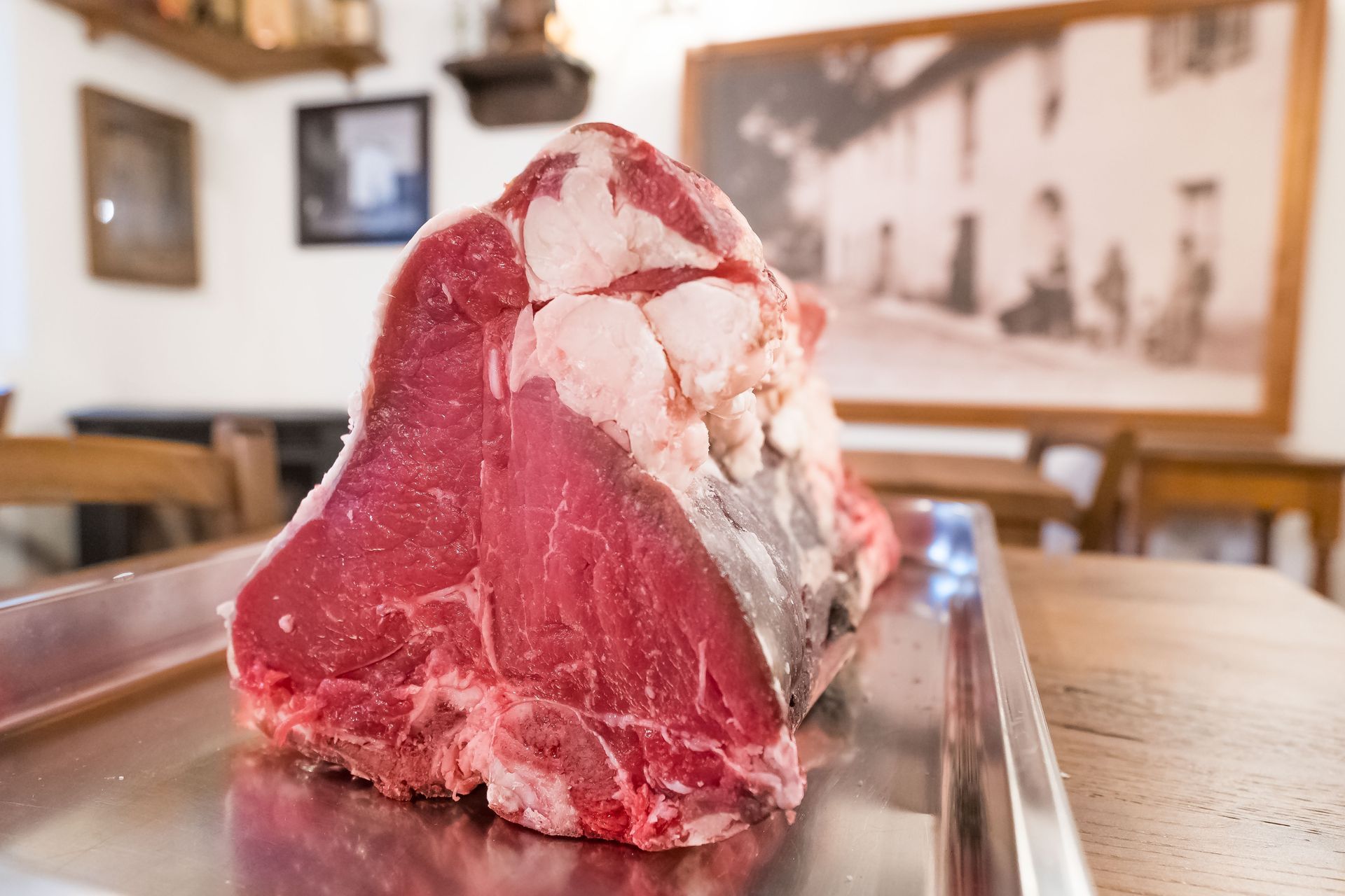 Una grande bistecca alla fiorentina cruda su un vassoio di metallo in un ristorante, con una foto incorniciata sul muro.
